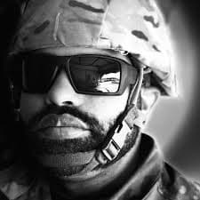Black and white close-up of a bearded soldier wearing a camouflage helmet and dark sunglasses.