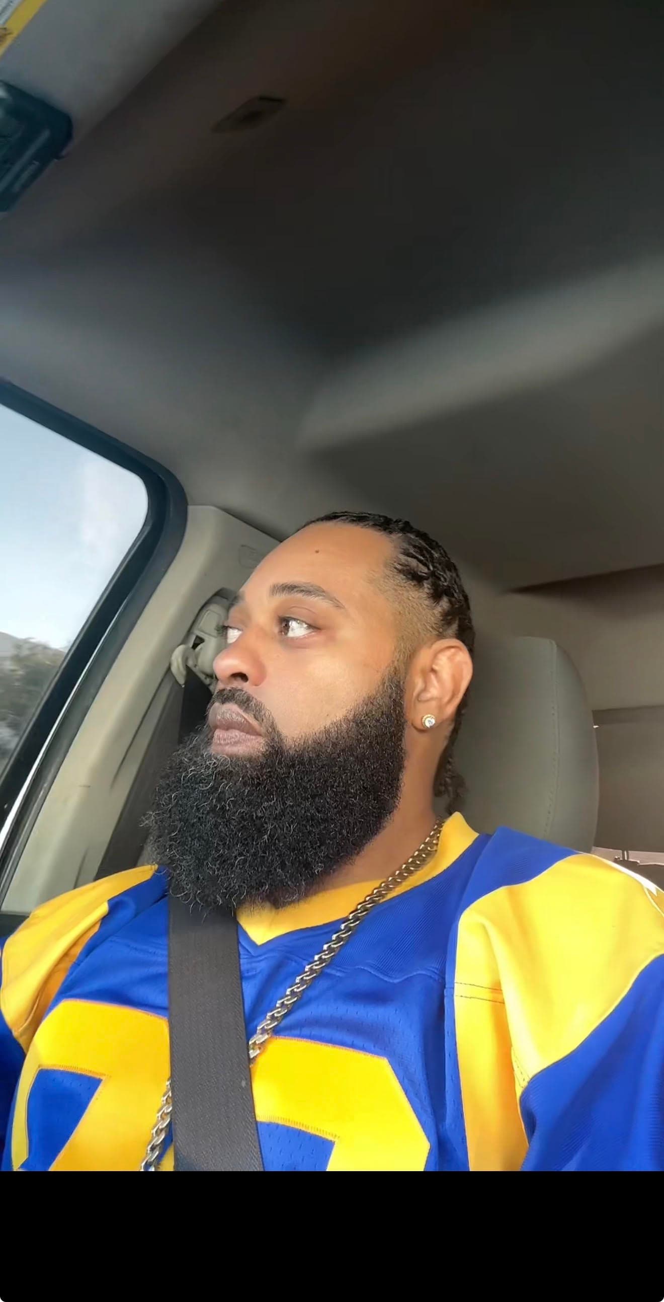 Bearded man with braided hair in a blue and yellow jersey looks out car window.